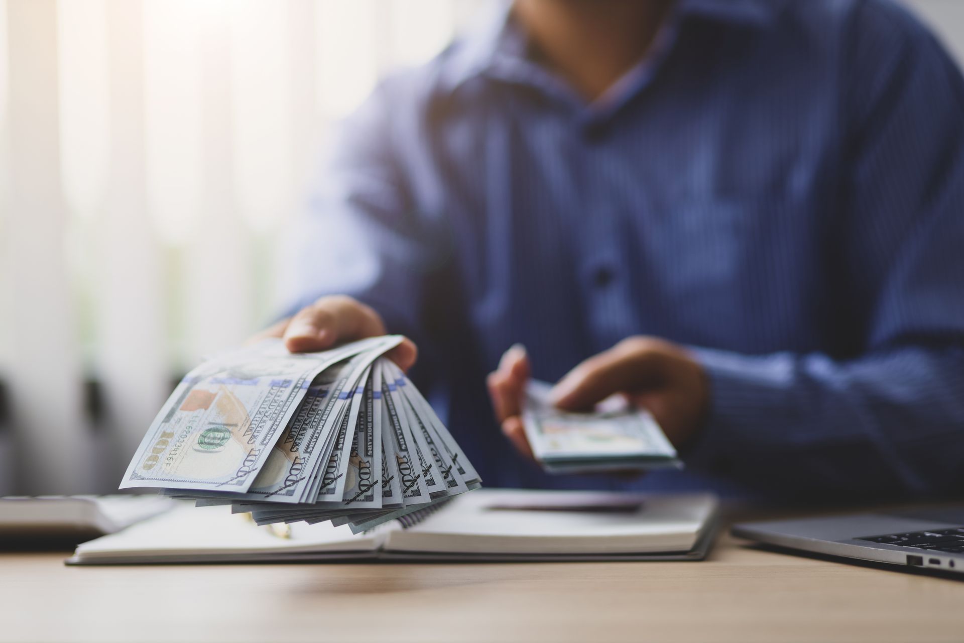 Person in dress shirt handing over a fanned stack of cash bills across an office desk.