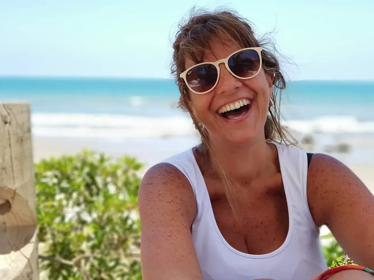 Mujer sonriente con gafas de sol en una playa soleada con el océano de fondo.