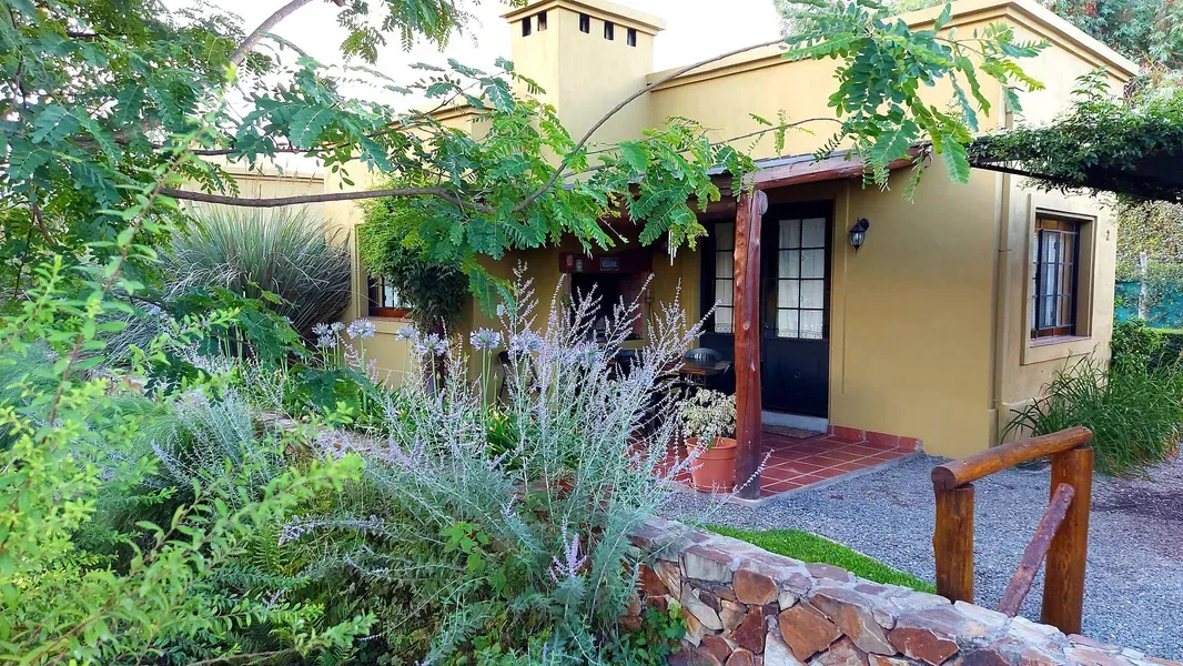Vista frontal de una pequeña casa de color beige con una puerta oscura, rodeada de exuberantes plantas verdes y un camino de ladrillos.