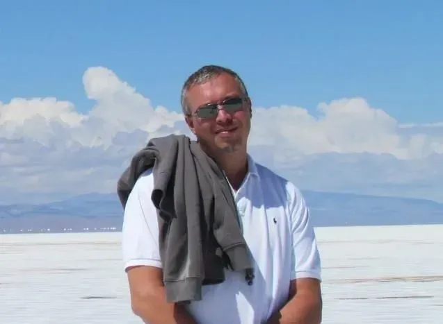 Hombre de pie con camisa blanca y chaqueta gris sobre el hombro, con gafas de sol, junto a una brillante salina bajo el cielo azul.