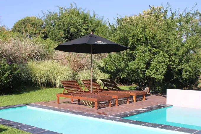 Tumbonas junto a la piscina y una sombrilla negra sobre una tarima de madera junto a una piscina turquesa, con una exuberante vegetación al fondo.