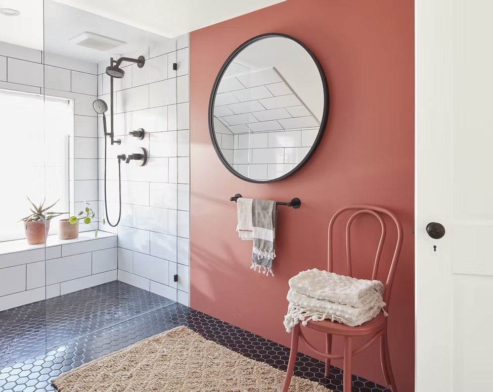 Stylish bathroom with white tile shower, pink accent wall, round mirror, and woven chair.