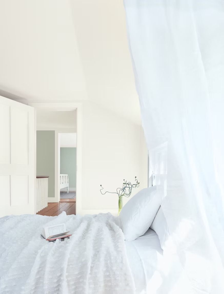 Bright white bedroom with a neatly made bed, sheer curtains, and an open doorway to a hallway.