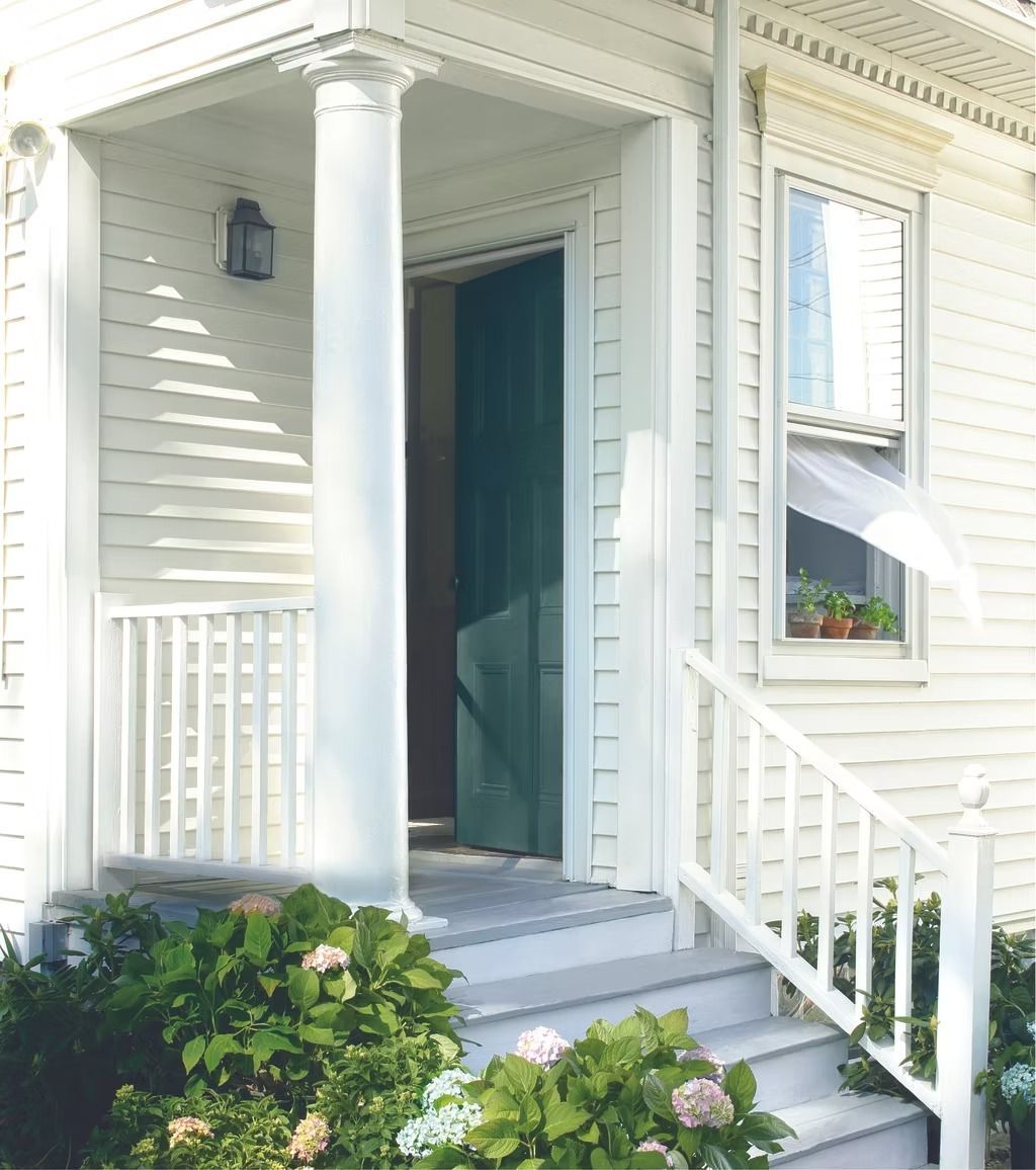 White house front porch with columns, open blue door, and steps beside flowers and greenery