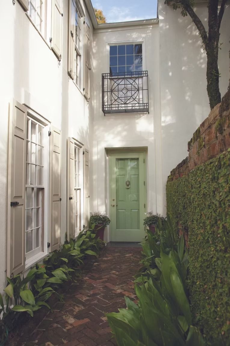 Narrow courtyard with white walls, green door, balcony, and brick path lined with plants