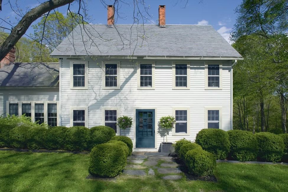 White two-story house with gray roof, blue door, and neatly trimmed hedges in a sunny green yard