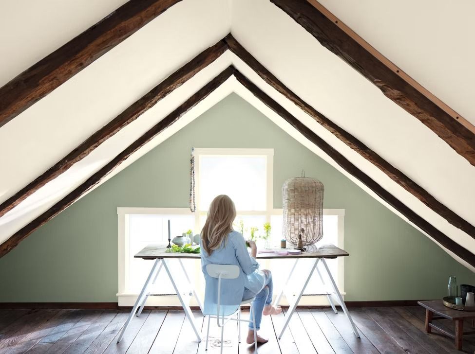 Person seated at a desk in an attic workspace with green walls and exposed wooden beams, facing a bright window