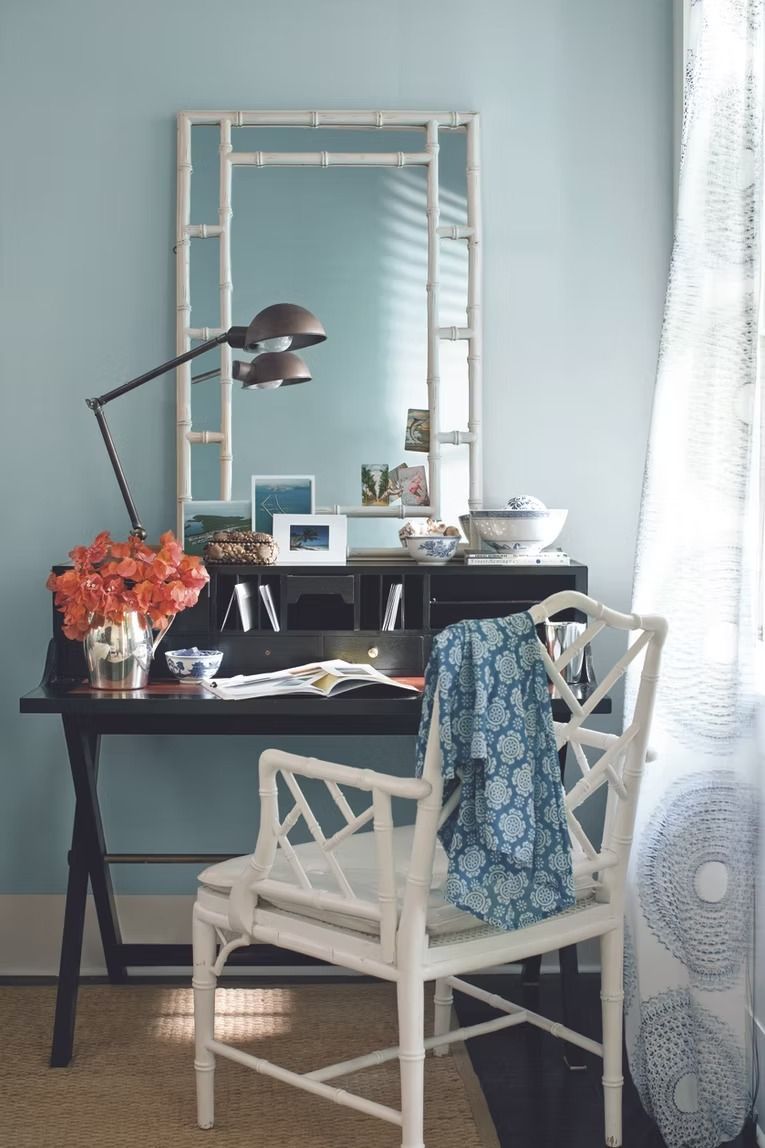 Cozy desk nook with black table, white chair, mirror, flowers, and blue curtain by the window