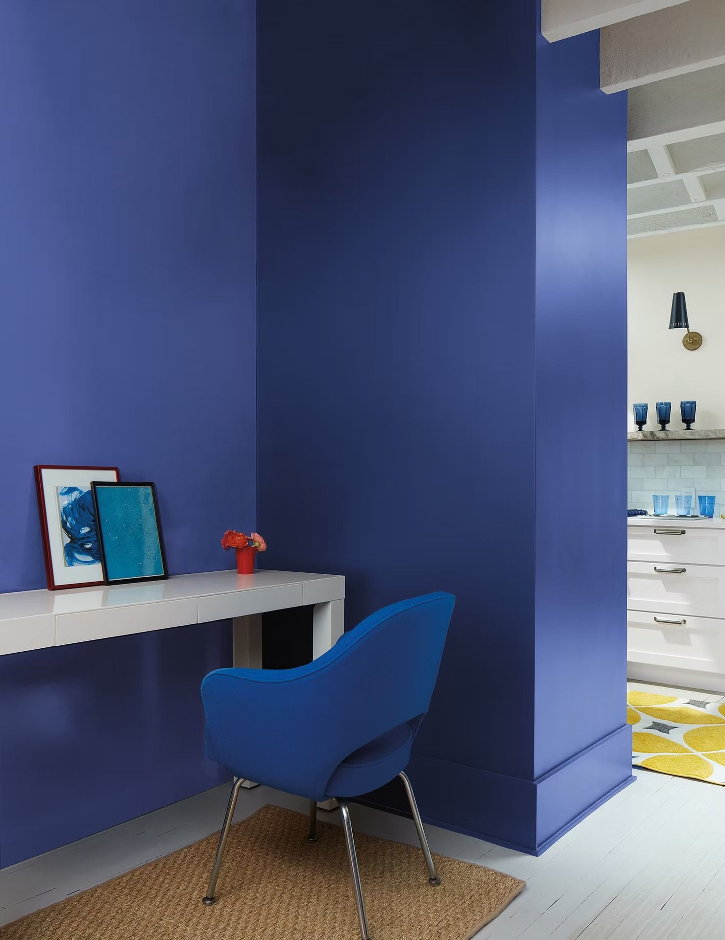 Blue chair and desk in a modern blue room with framed art and a view into a bright kitchen.