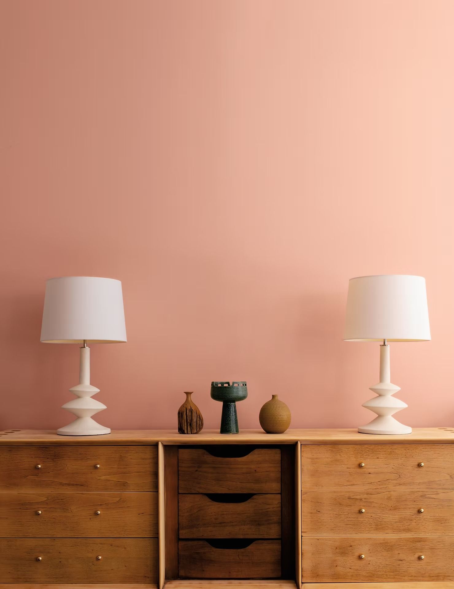 Two white table lamps on a wooden dresser against a pink wall, with small decor pieces in the center.