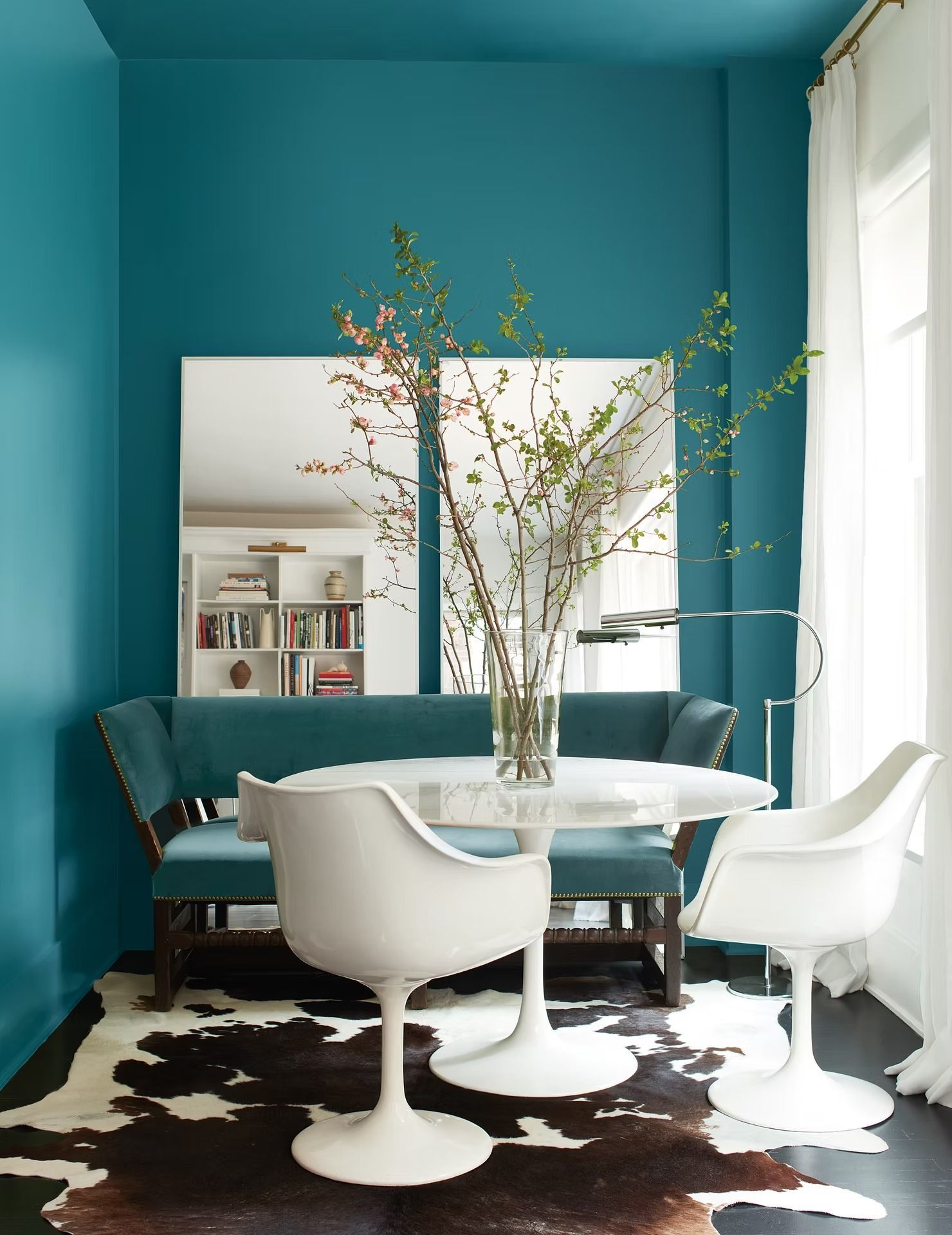 Teal dining room with round table, white chairs, mirror, and tall vase of branches