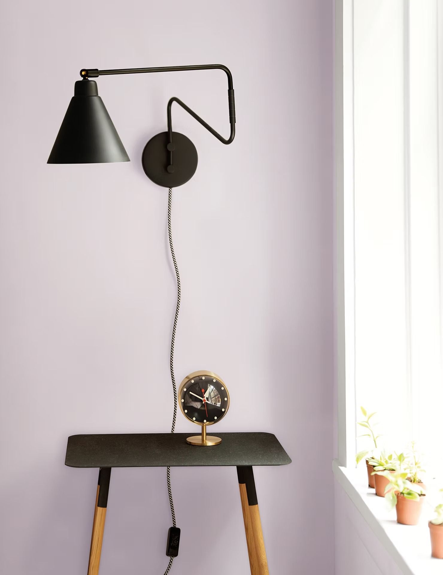 Black wall lamp over small wooden table with a round mirror, beside a bright window and potted plants.