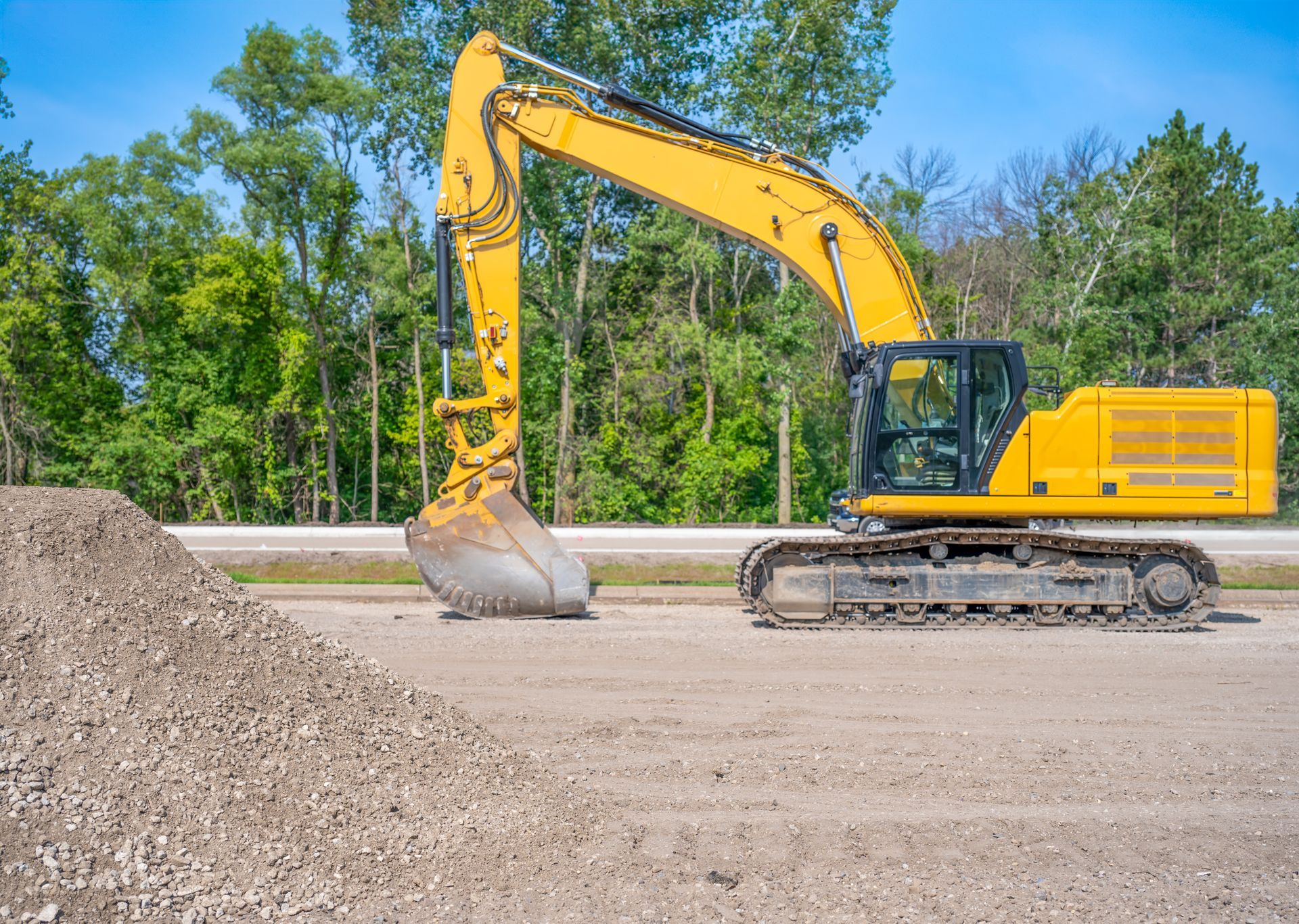 A yellow excavator parked on a dirt lot next to a pile of gravel with trees and a clear blue sky in the background.