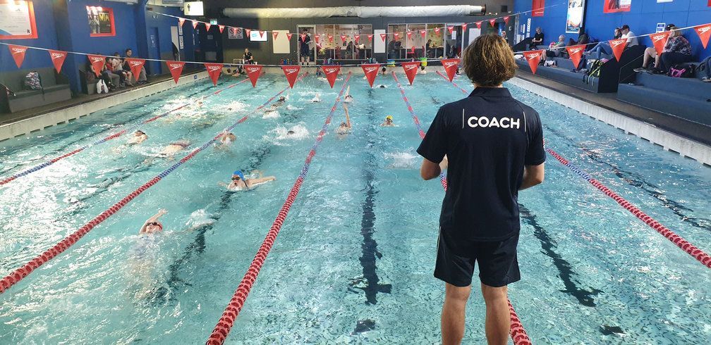 A coach is standing in front of a swimming pool watching people swim.