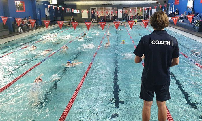 A coach is standing in front of a swimming pool watching people swim.