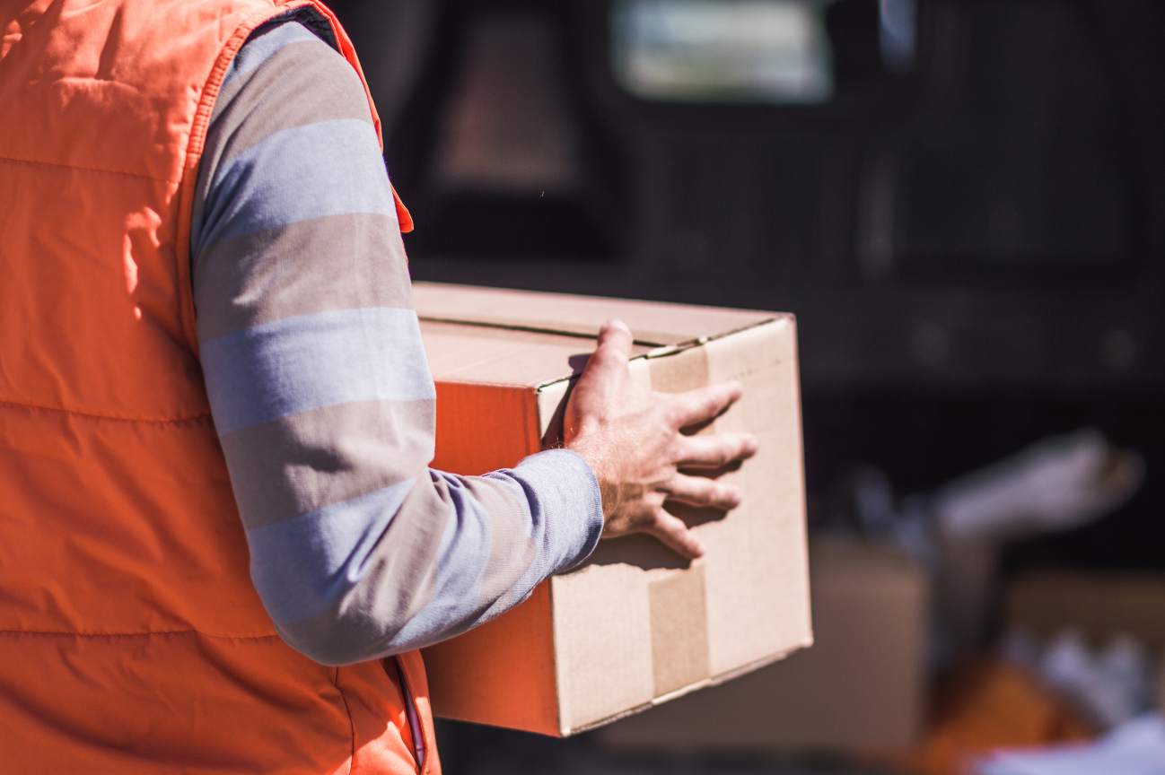 A man in an orange vest is carrying a cardboard box.