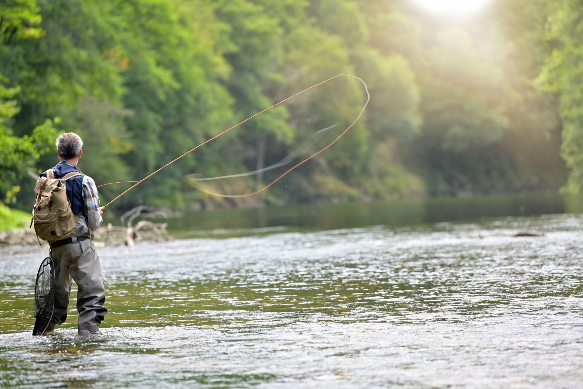 Angler standing in a river casting a fishing rod, with green trees and sunlight in the background