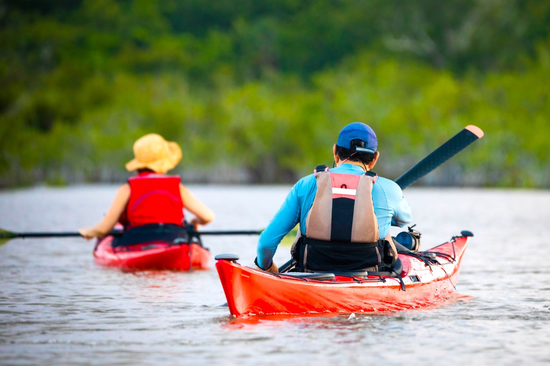 Two kayakers in red kayaks on a calm lake, paddling away toward a green shoreline.