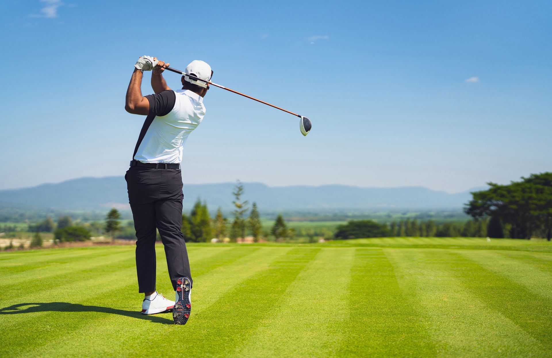 Golfer in white shirt swinging club on a green course with mountains in the background