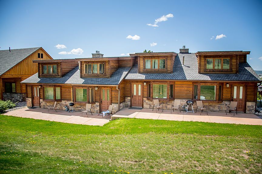 Wooden lodge-style house with dormer windows and patio seating on a sunny lawn under a blue sky