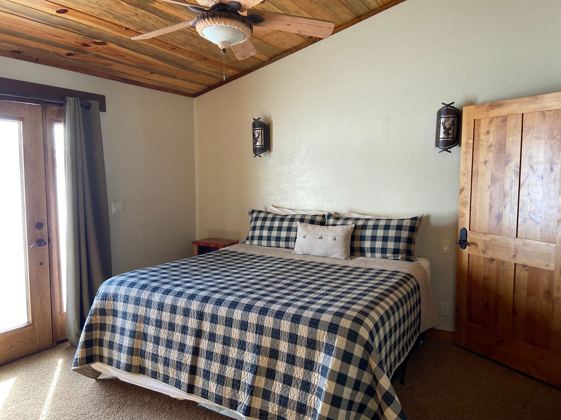 Rustic bedroom with plaid bedspread, wood ceiling, bedside lamps, and wooden door.