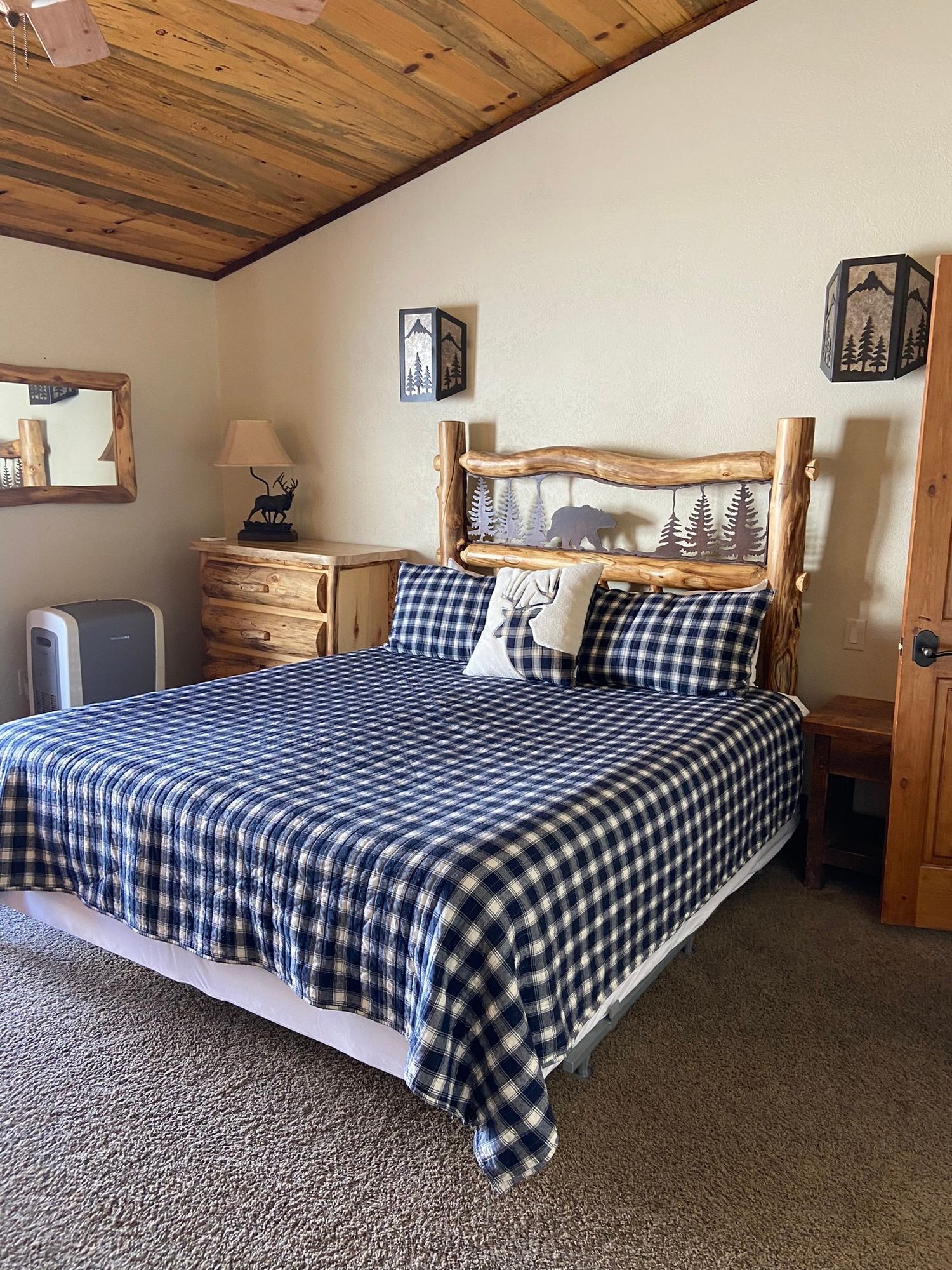 Cozy bedroom with a blue plaid bedspread, wooden furniture, and a slanted wood-paneled ceiling