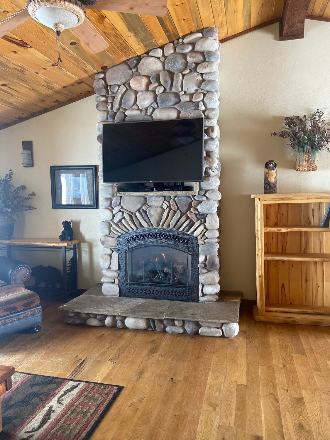 Rustic living room with stone fireplace, wall-mounted TV, wood ceiling, and hardwood floors