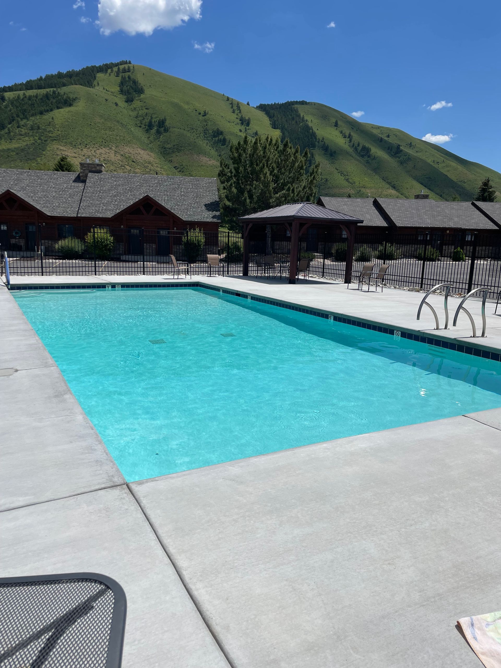 Outdoor pool with turquoise water, white deck, lounge chairs, and green hills under a blue sky.