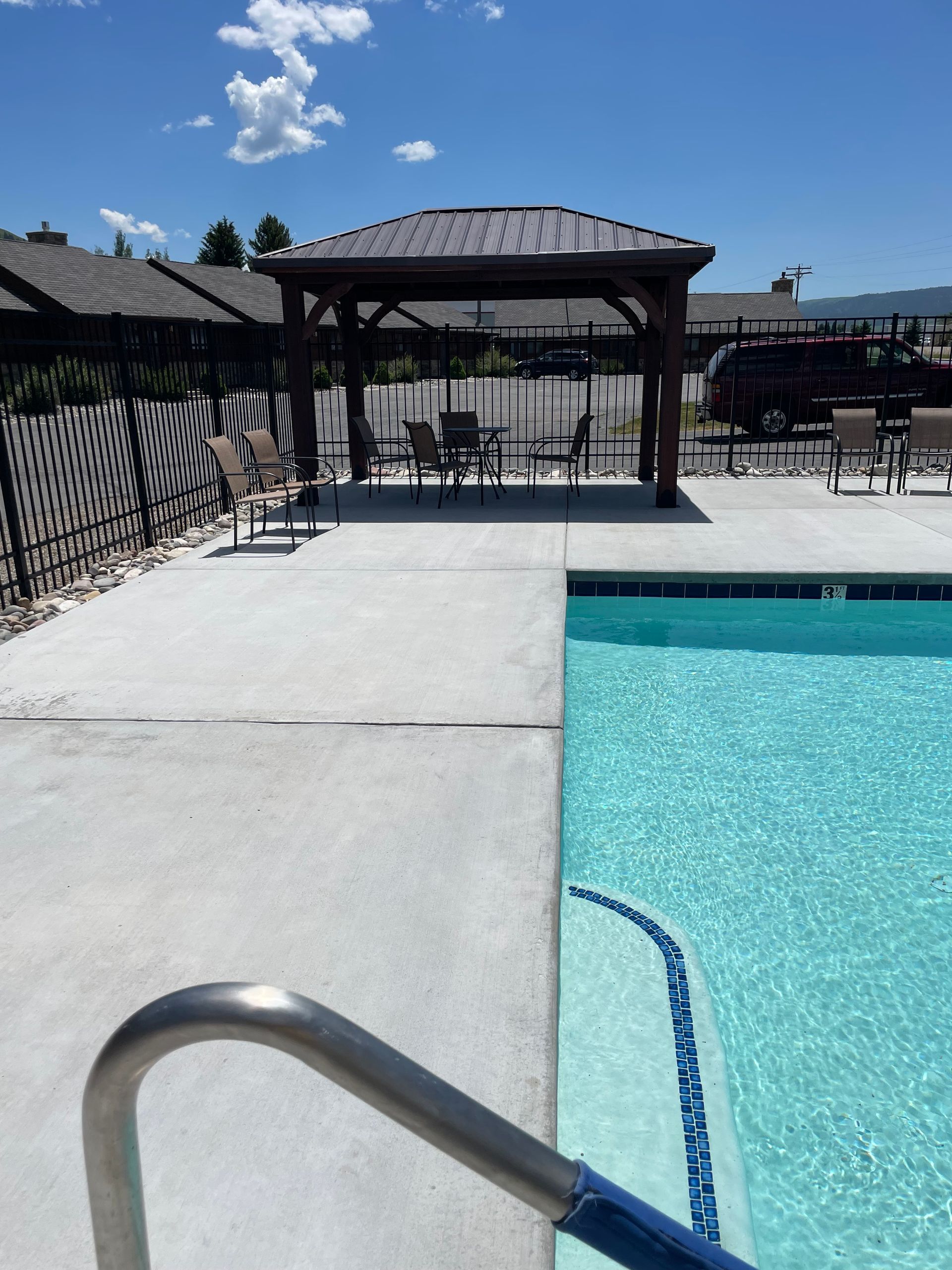 Outdoor pool with blue water, concrete deck, and shaded seating area under a pavilion on a sunny day