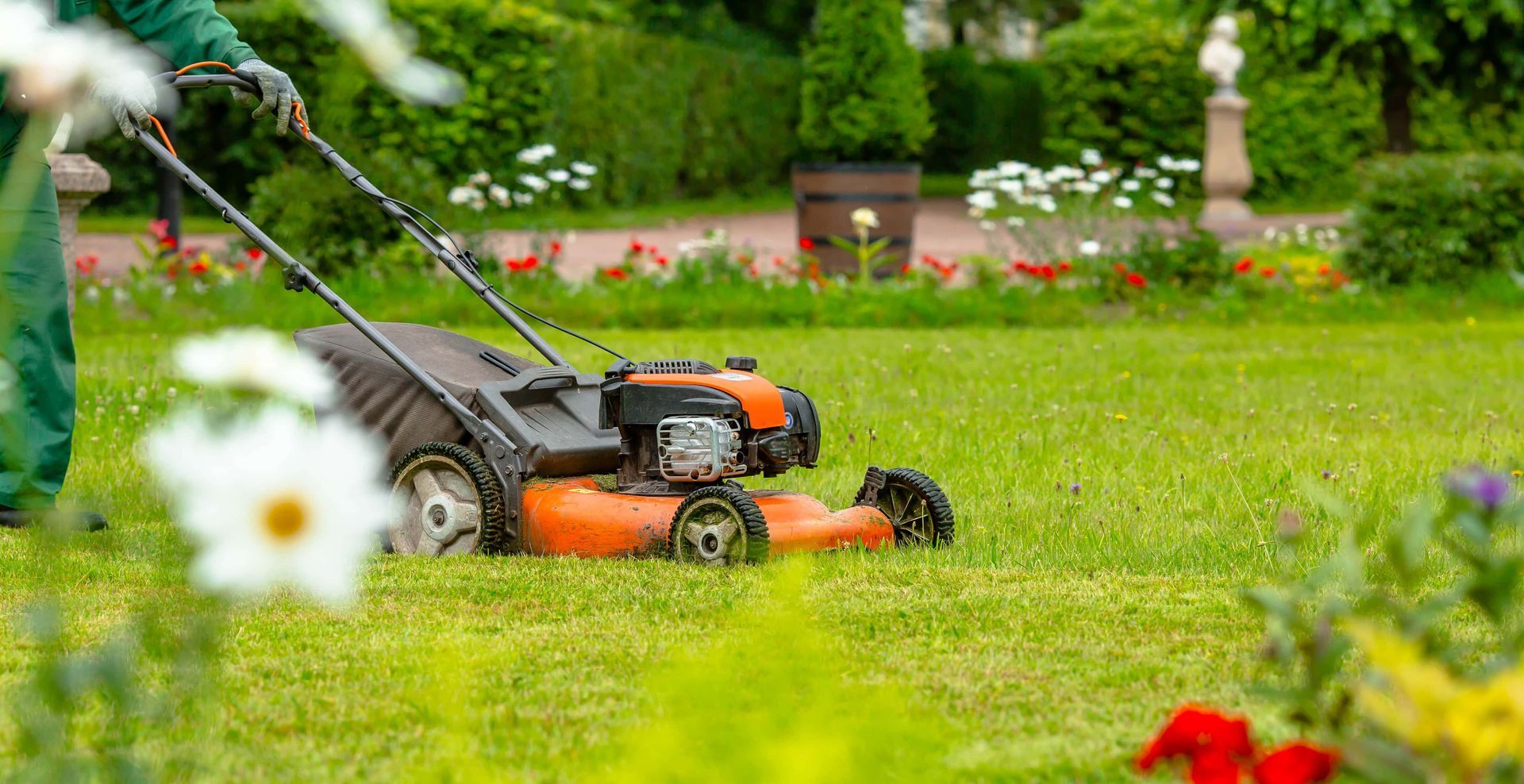 Person mowing a green lawn with an orange and black lawnmower in a garden.