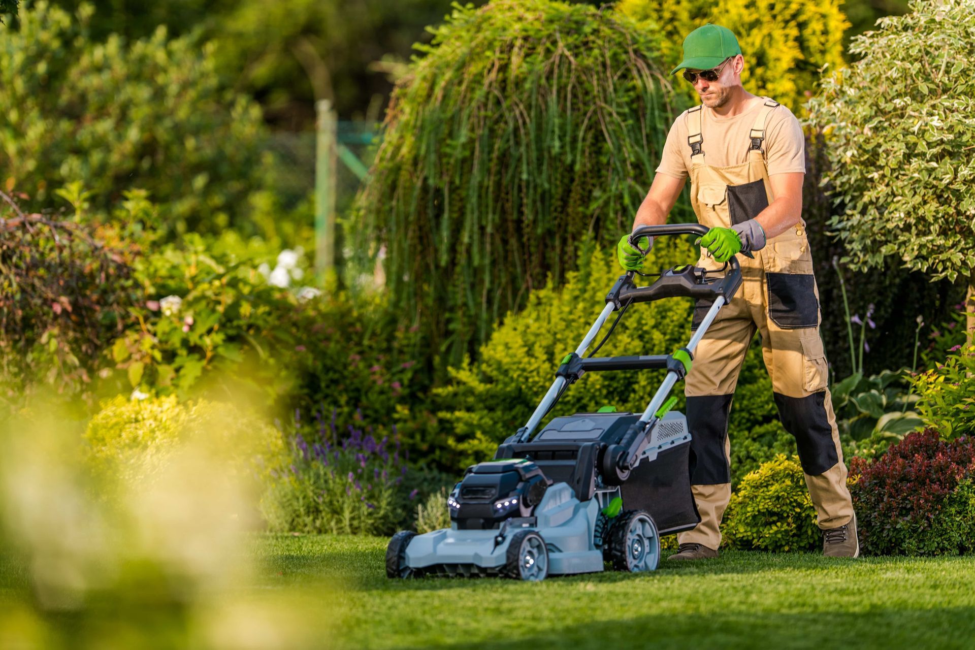 Man in green hat mowing a lush green lawn with an electric lawnmower in a garden.