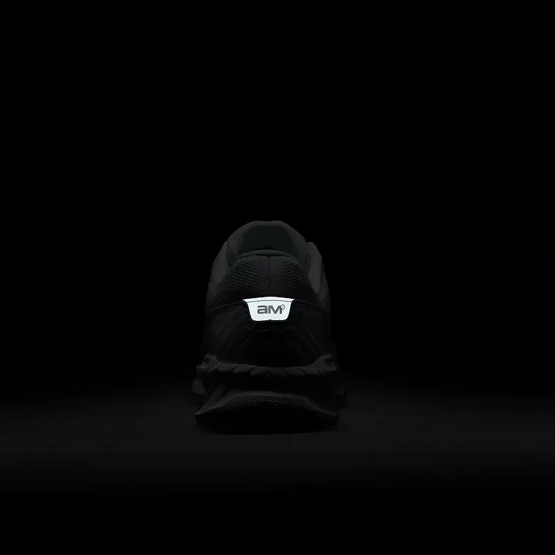 Black sneaker with a reflective logo, viewed from the heel, against a dark background.