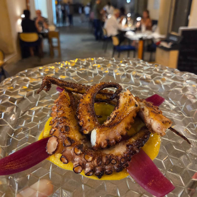 Polpo grigliato servito su un piatto con salsa gialla e guarnizioni viola in un ambiente da ristorante.