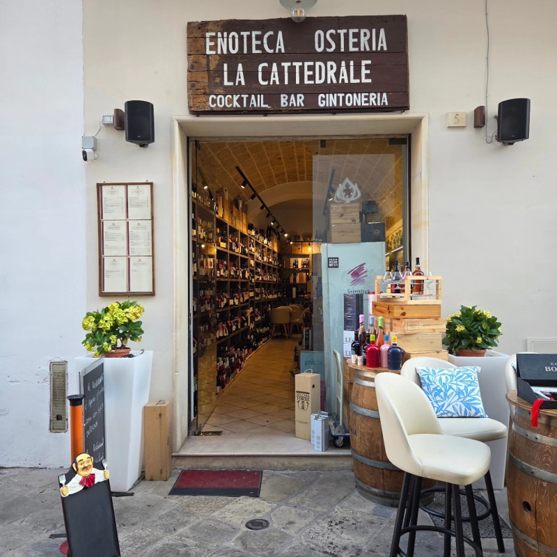 Esterno dell'Enoteca Osteria La Cattedrale, con porta d'ingresso aperta, scaffali per il vino e tavolini all'aperto.