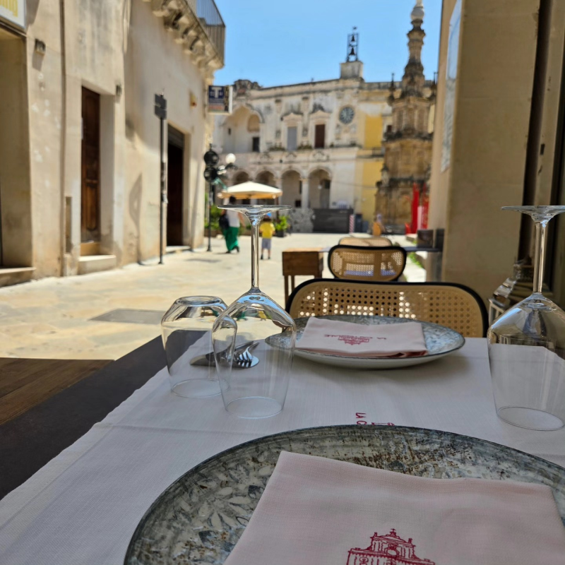 Tavolino da caffè all'aperto con bicchieri e piatti, affacciato su una piazza storica soleggiata con edifici riccamente decorati.