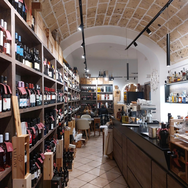 Interno di un'enoteca con scaffali pieni di bottiglie, pavimento piastrellato e soffitto a volta in mattoni.