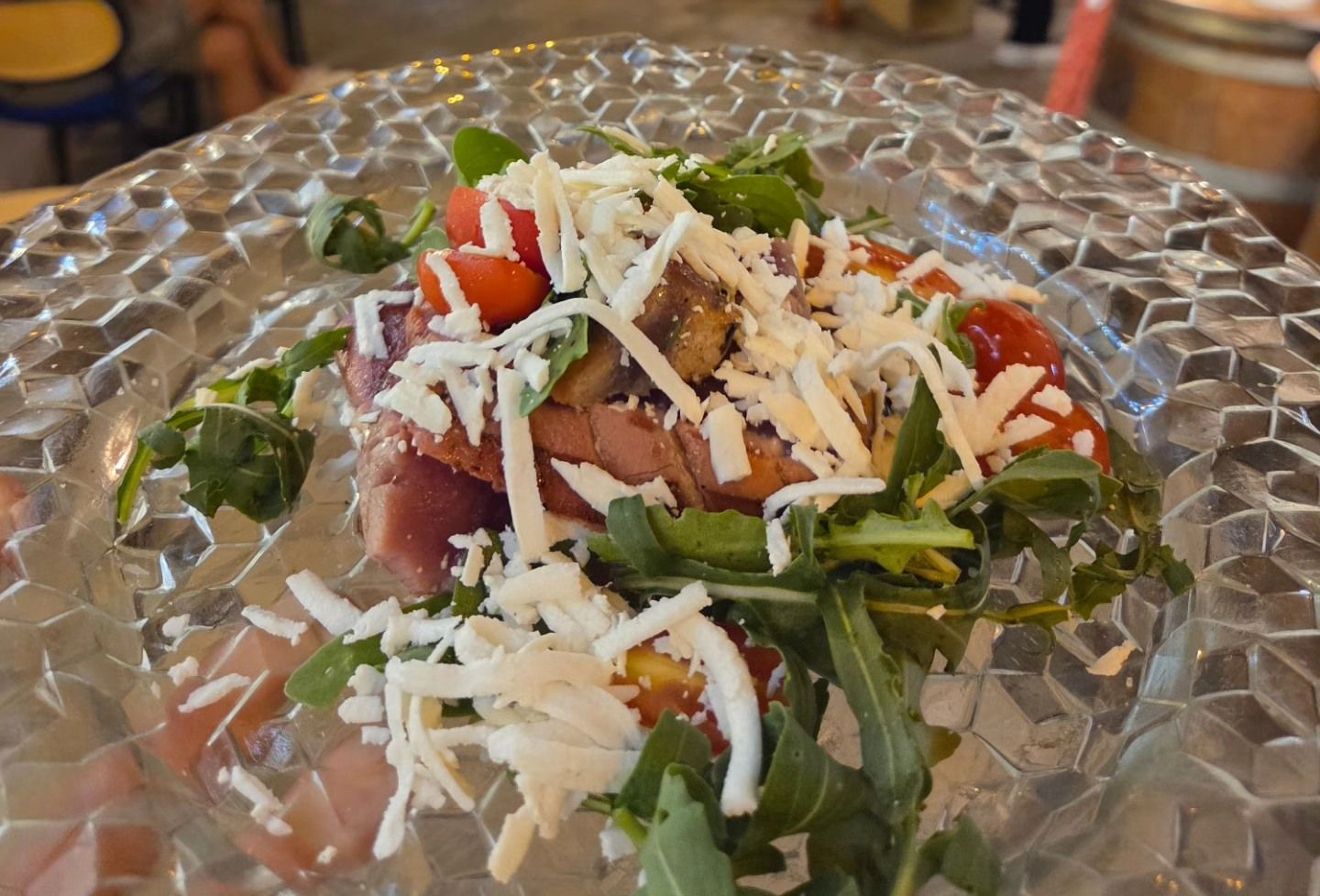 Insalata con verdure a foglia verde, pomodori, affettati e formaggio grattugiato su un piatto di vetro testurizzato.