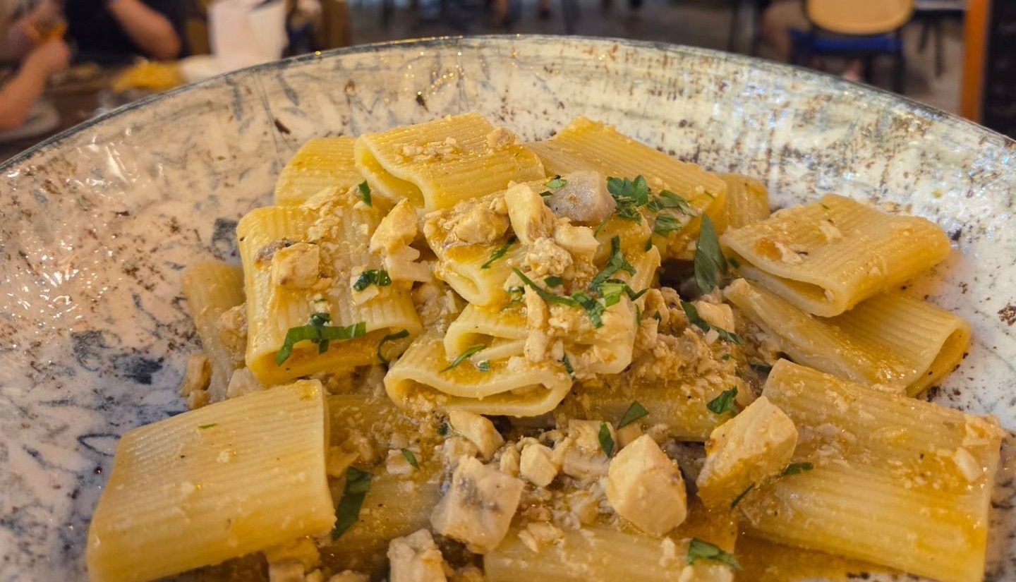 Rigatoni in salsa cremosa, conditi con formaggio grattugiato ed erbe aromatiche, serviti in una ciotola rustica.