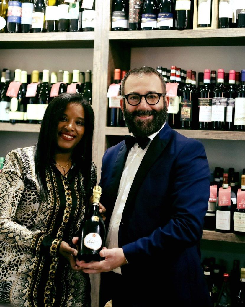 Due persone sorridenti che tengono in mano una bottiglia di vino davanti a degli scaffali pieni di bottiglie di vino.