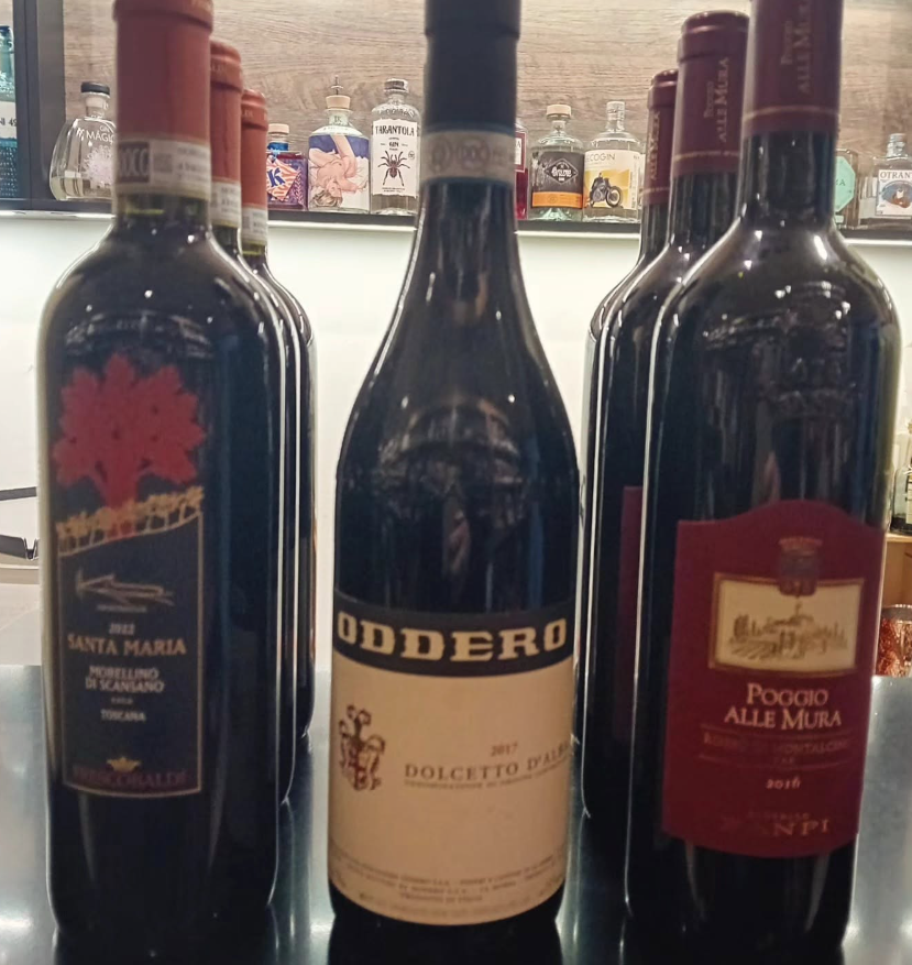 Quattro bottiglie di vino rosso scuro allineate su un bancone, con le etichette delle bottiglie davanti a uno sfondo sfocato.
