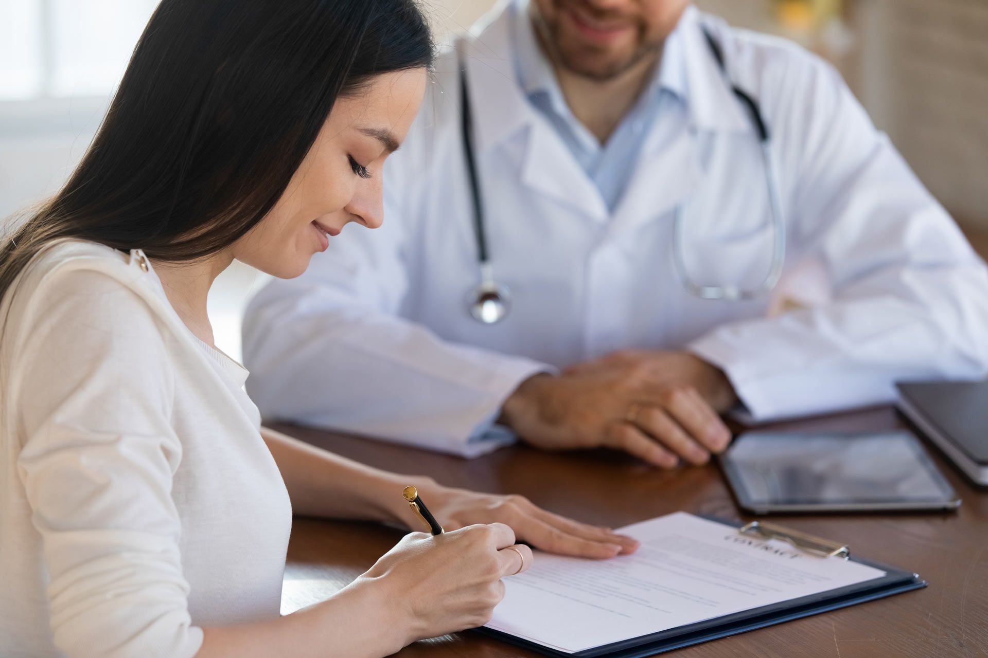 A woman is sitting at a table with a doctor and signing a document