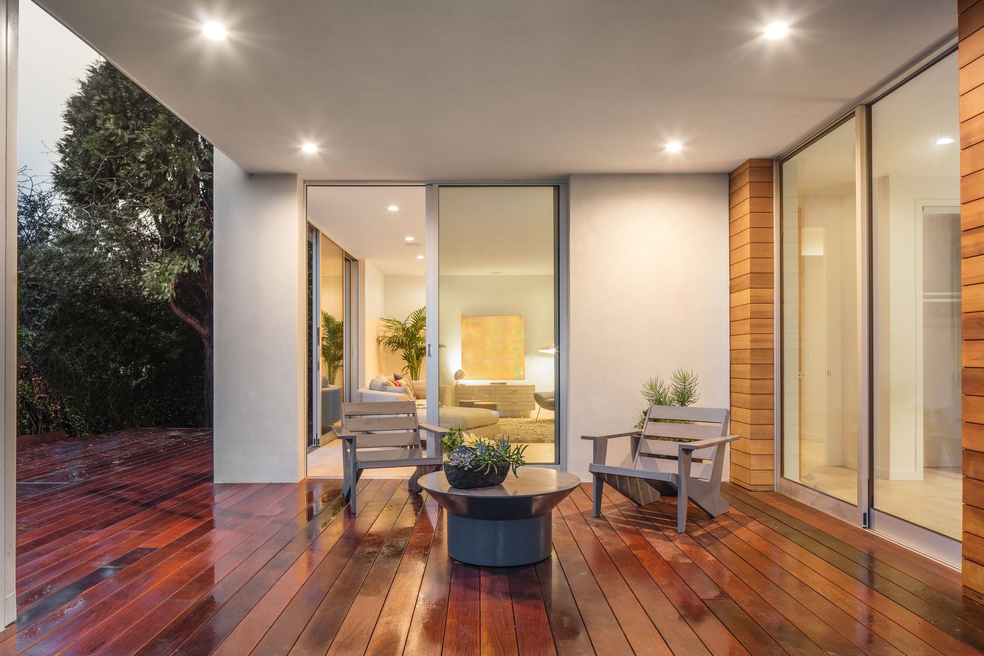 Modern patio with wooden deck, outdoor seating, and warm interior lighting beside large glass doors