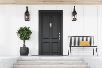 Black front door with lanterns, potted tree, and striped bench on a white porch.