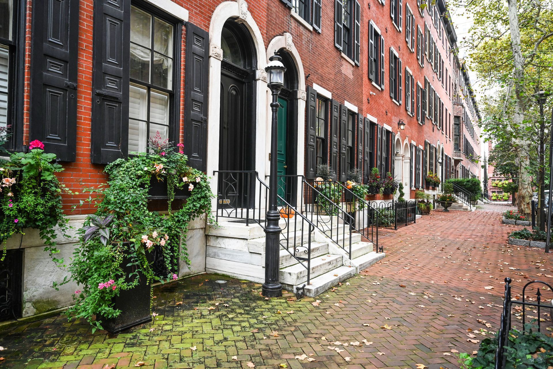 Red brick rowhouses with black shutters and flower boxes along a leafy cobblestone street