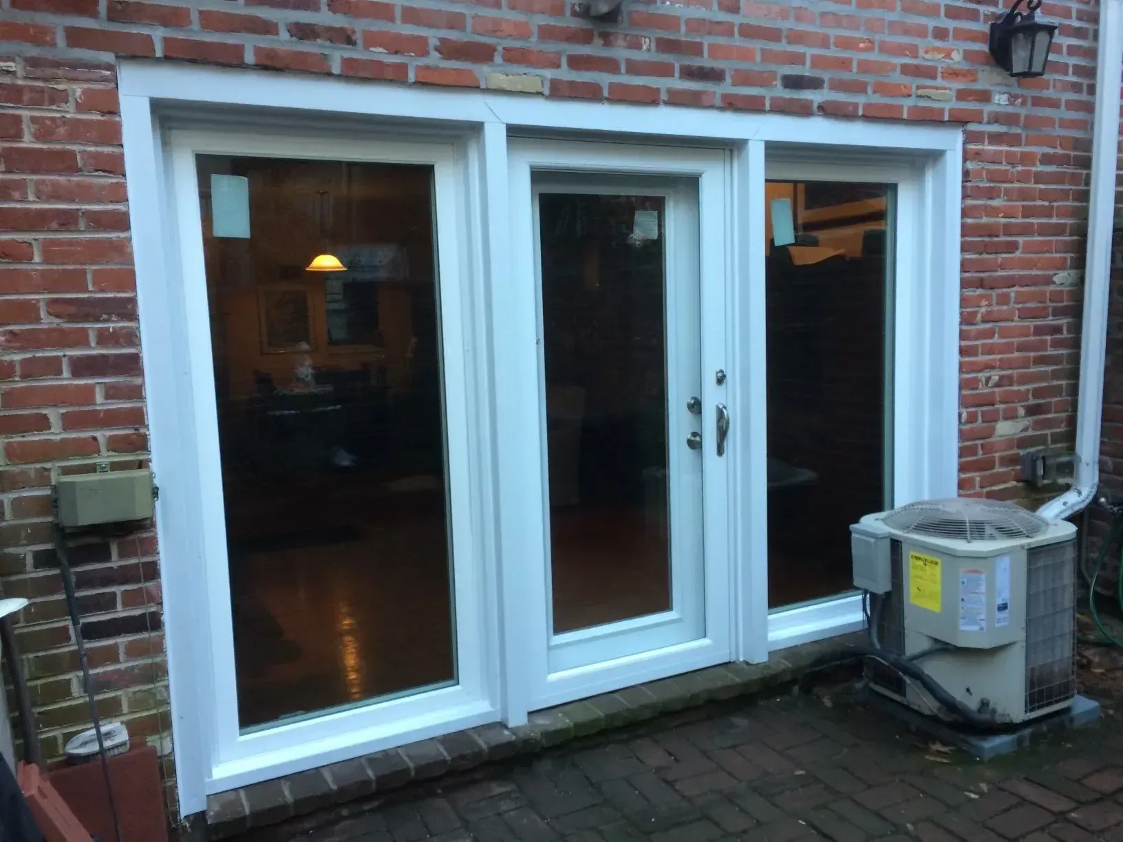 White-framed glass patio doors in a brick wall, with a trash bin on the right and warm light inside.