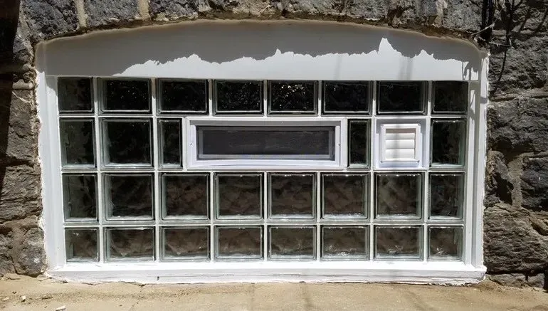 Stone wall with a small white-framed basement window and vent grate above a concrete sill