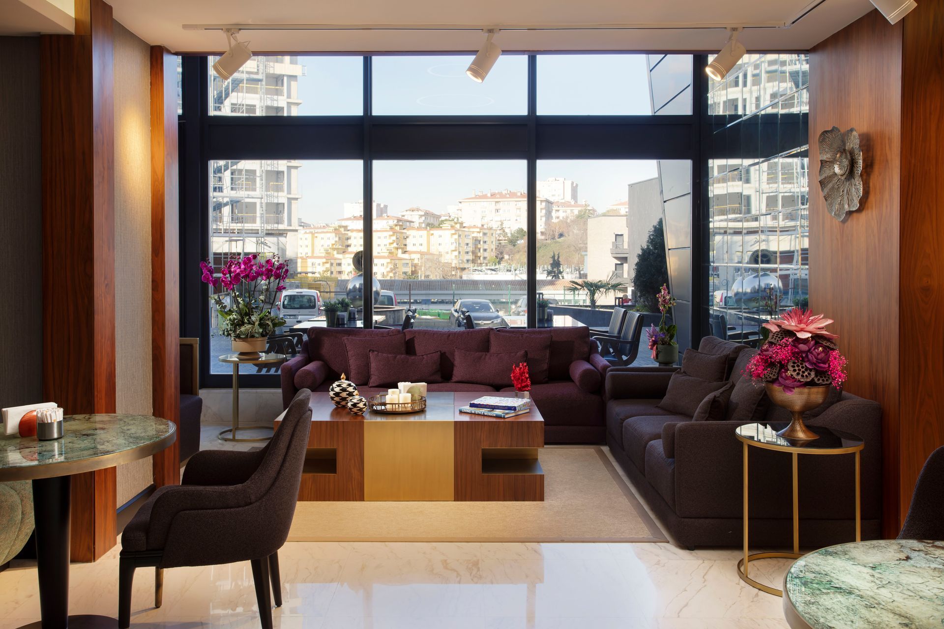 Geniş pencereleri, koyu renk kanepeleri, sehpası ve gün batımında şehir manzarasıyla modern otel lobisi.