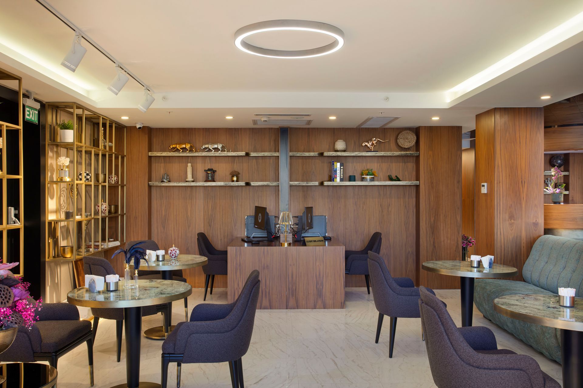 Masalar, sandalyeler, ahşap raflar ve resepsiyonun bulunduğu, sıcak aydınlatmalı modern otel lobisi.