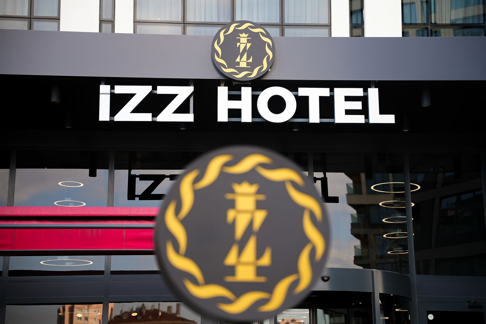 Bir dükkanın vitrininin üzerinde, altın defne yaprağı logosu ve kırmızı yatay çizgilerle süslenmiş IZZ Otel tabelası.