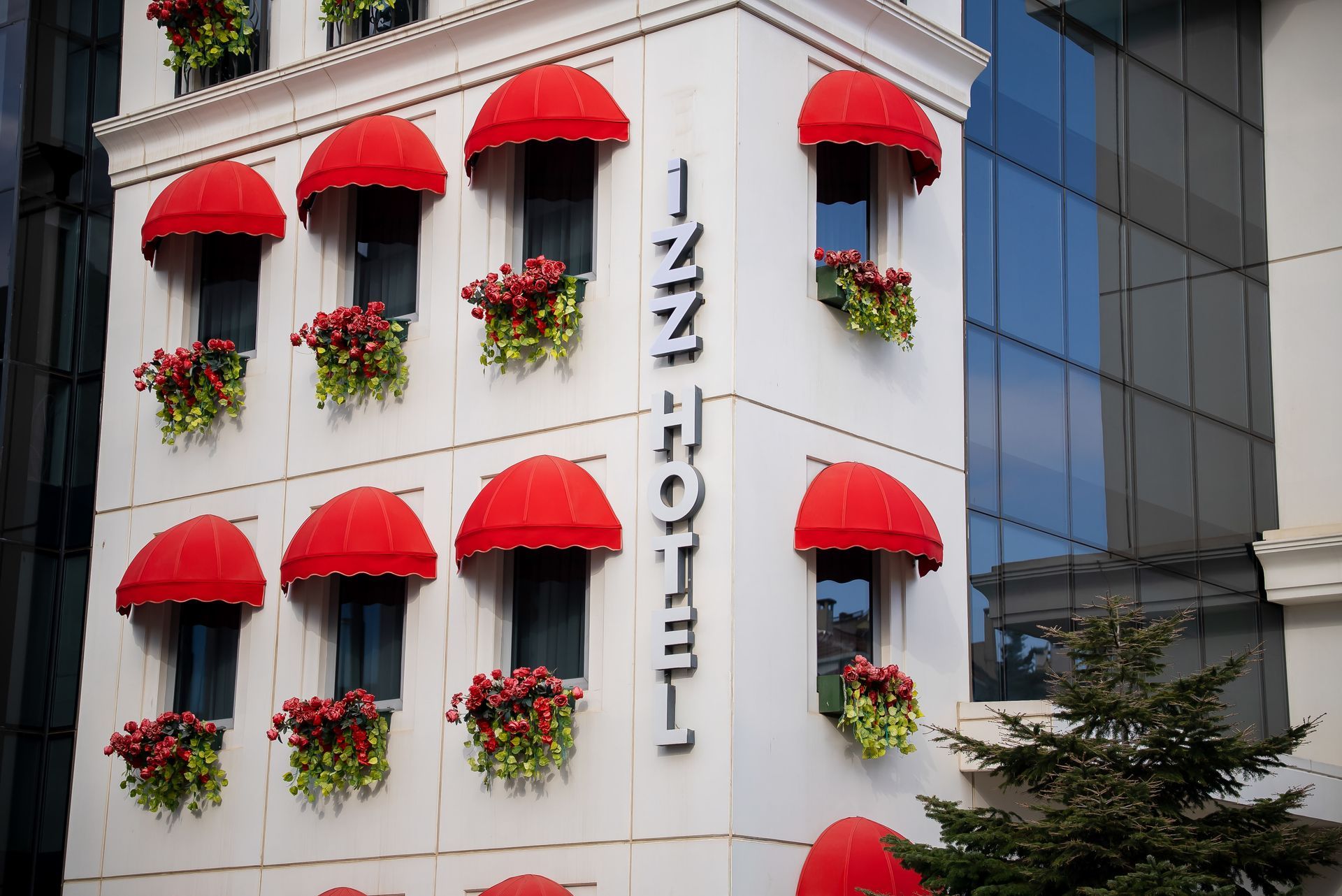 Kırmızı tenteler ve çiçek kutularıyla süslenmiş, dikey otel tabelası bulunan beyaz bina cephesi.